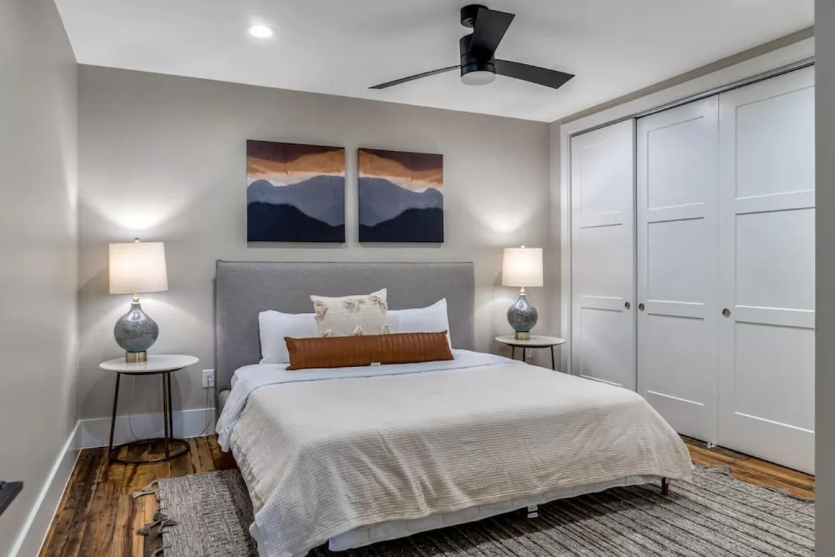 This image features a king-sized bed adorned with decorative pillows, flanked by nightstands each with a lamp for soft lighting. Above the bed hangs tasteful artwork, and the overall color palette and furnishing choices create a calming, upscale atmosphere—ideal for restful nights or relaxed reading.