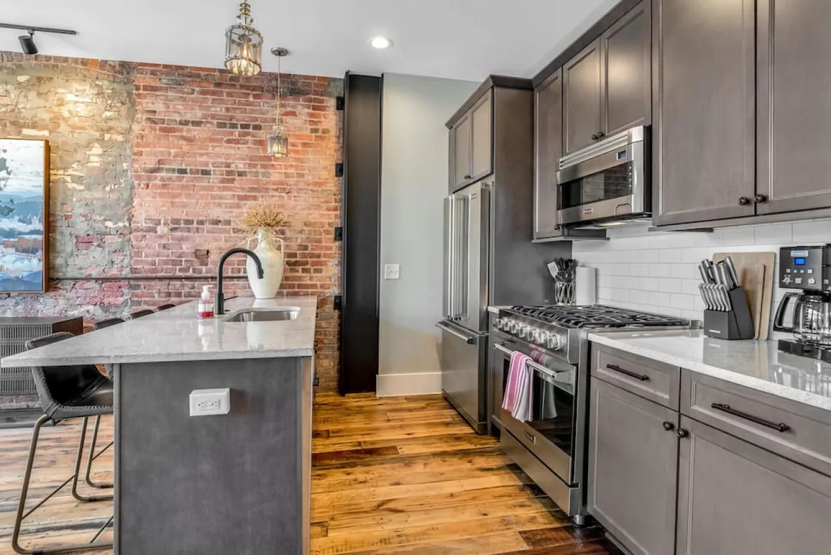 A stylish modern kitchen with granite countertops, stainless steel appliances, exposed brick wall, and elegant pendant lighting. The image shows a cooking area ready for meal prep, with a breakfast bar inviting for coffee or casual dining. The blend of modern finishes and warm textures creates an inviting, chef-inspired atmosphere.