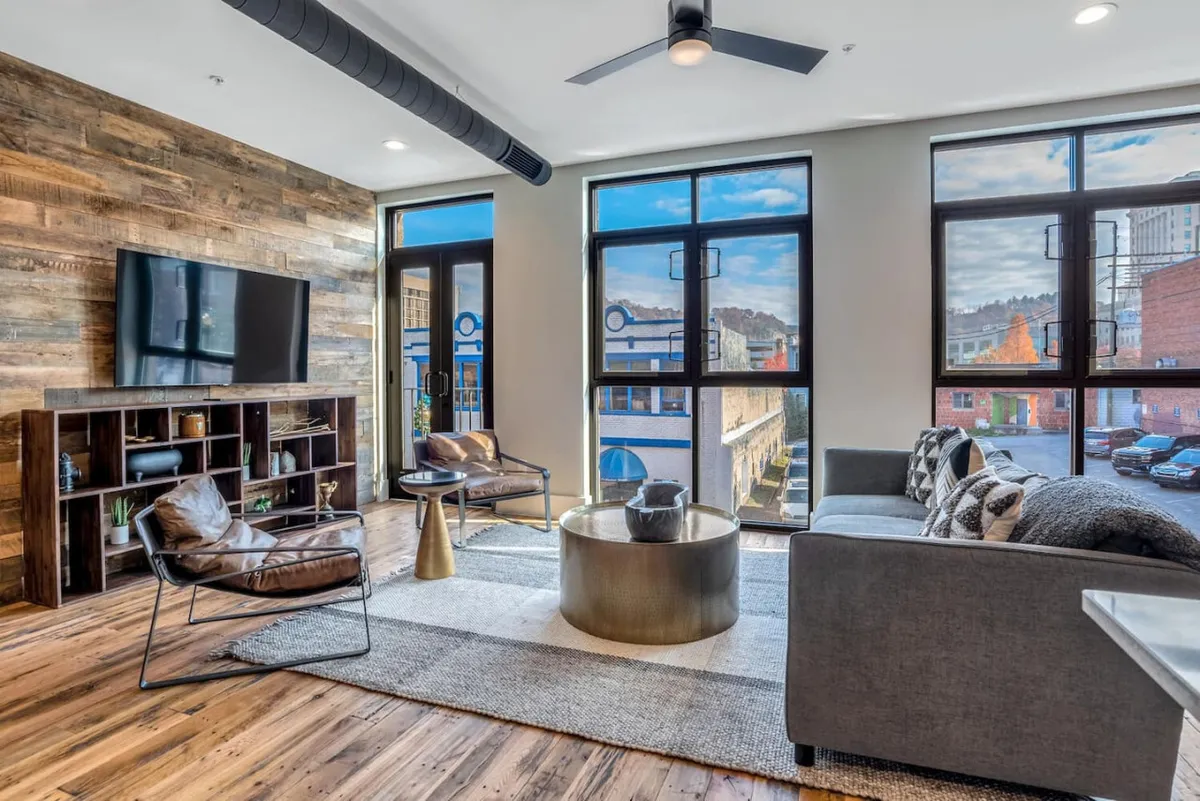 A luminous living area featuring a wood feature wall, expansive large windows that flood the space with natural light, and a mix of modern furniture. The coffee table and seating arrangement create a welcoming space for socializing or relaxing while observing the city outside.