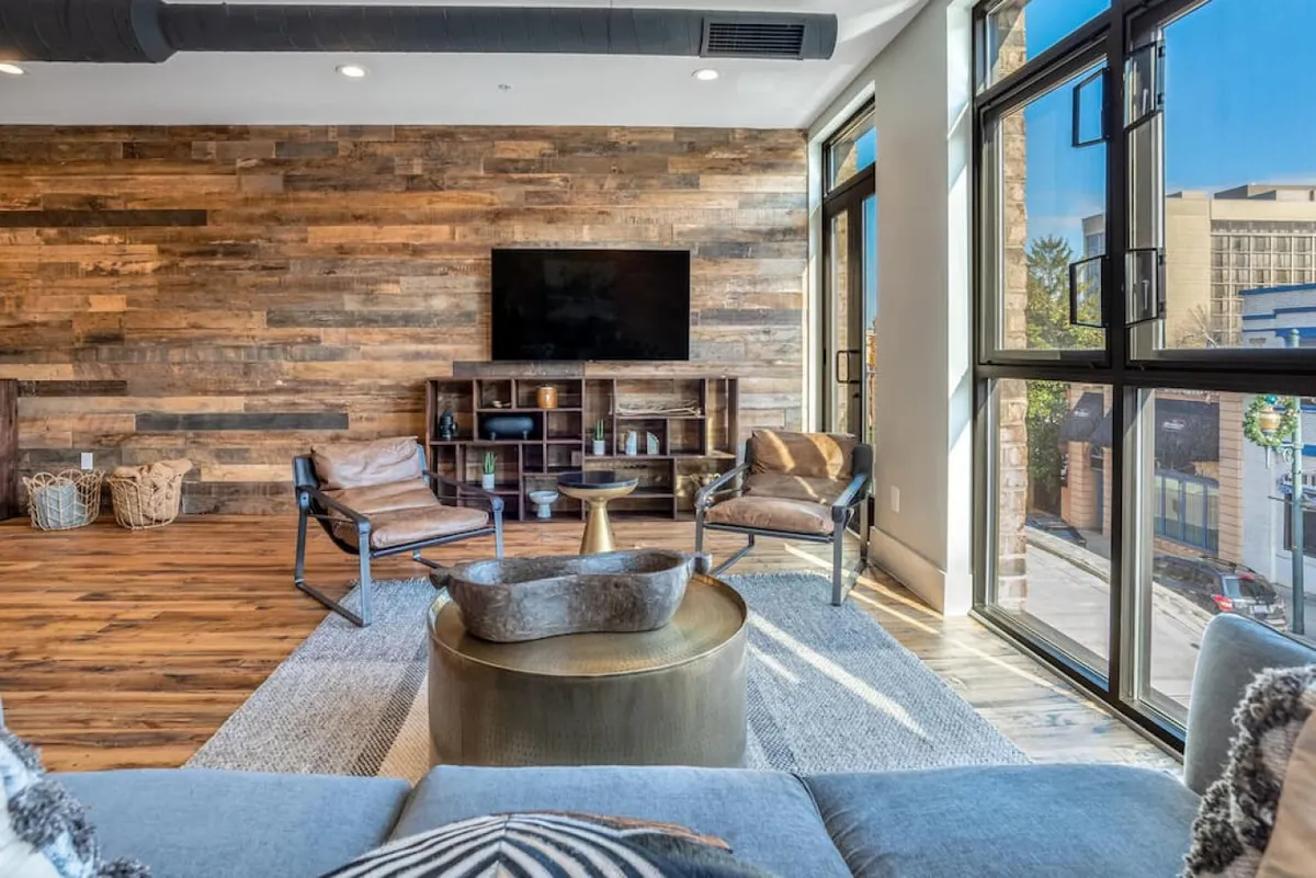 This image showcases a cozy, stylish seating area with a wooden accent wall, TV, coffee table, and soft natural light streaming in. The modern decor and comfortable furnishings create a welcoming environment ideal for relaxing or socializing after a day in Asheville.