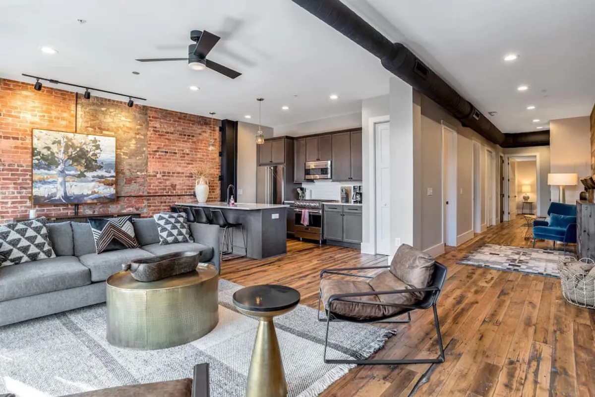 A stylish open-concept space defined by an exposed brick wall, a modern kitchen island, comfortable seating, and an eye-catching art piece. The area exudes warmth and character, with vintage accents and spaces for both entertaining and quiet relaxation.