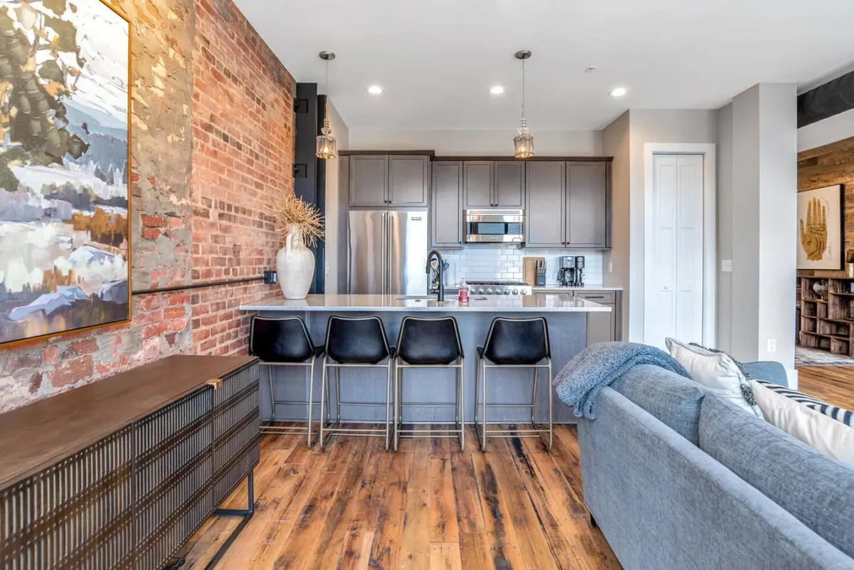 A contemporary kitchen scene featuring an exposed brick wall, stylish bar stools, modern cabinetry, and an art piece on the wall. The kitchen layout flows into a casual dining area, emphasizing spaces for both cooking and connection.