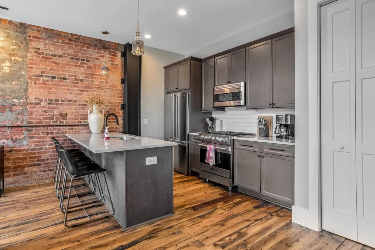 An inviting kitchen with an island and bar stools, exposed brick wall, and stainless steel appliances. This layout encourages conversation and collective meal preparation, embodying the open, interactive vibe mentioned in the narrative.