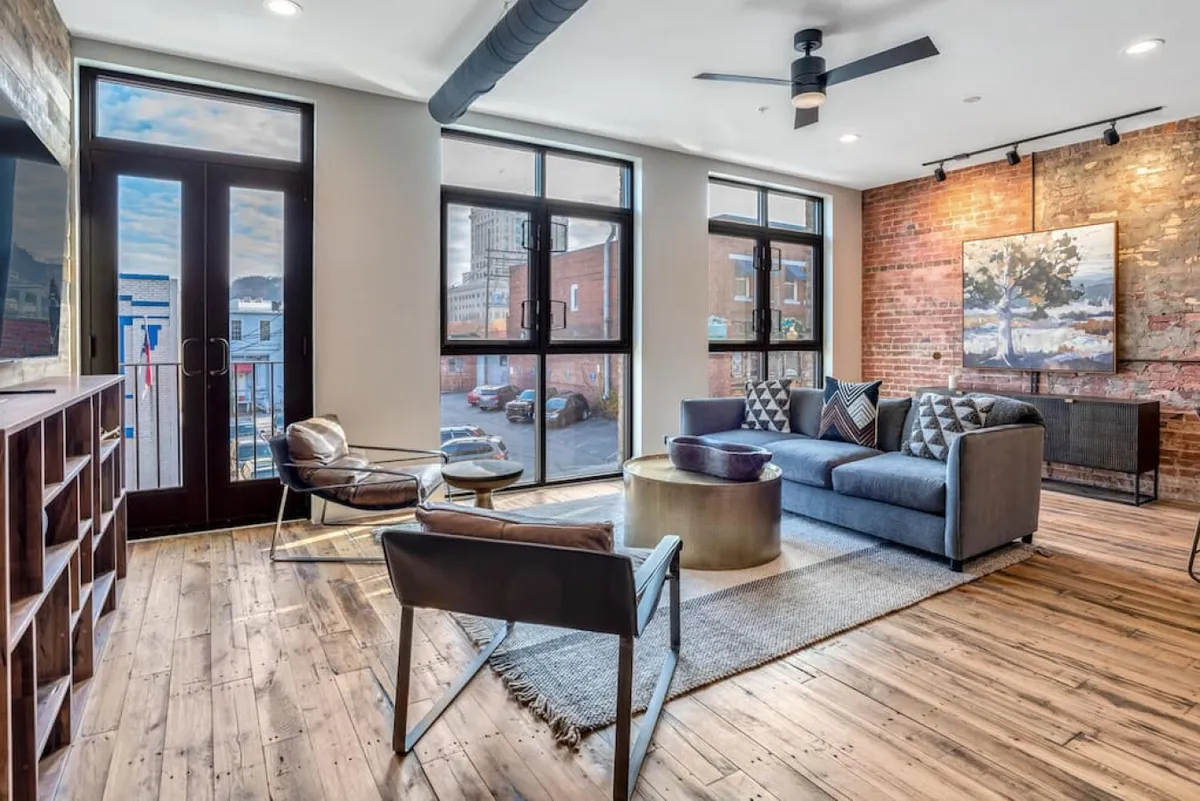 A stylish living room blends exposed brick walls, large sunlit windows with sweeping city views, a plush sectional sofa, a chic metallic coffee table, and modern art, creating a space that feels both historic and contemporary.