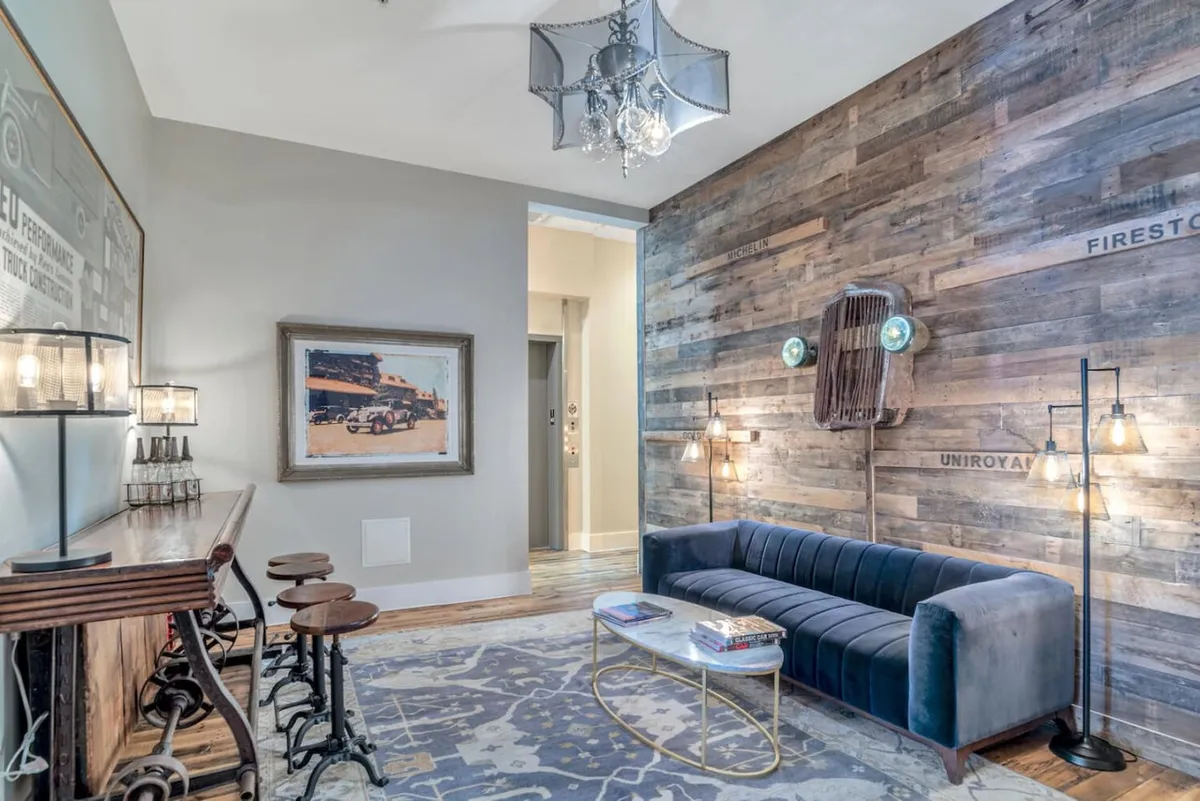 A stylish living area featuring a striking wood accent wall, plush blue velvet sofa, elegant chandelier, and modern artwork. Bar stools line the open-plan space, inviting guests to relax or socialize. Natural light and a blend of textures create a contemporary yet comfortable atmosphere.